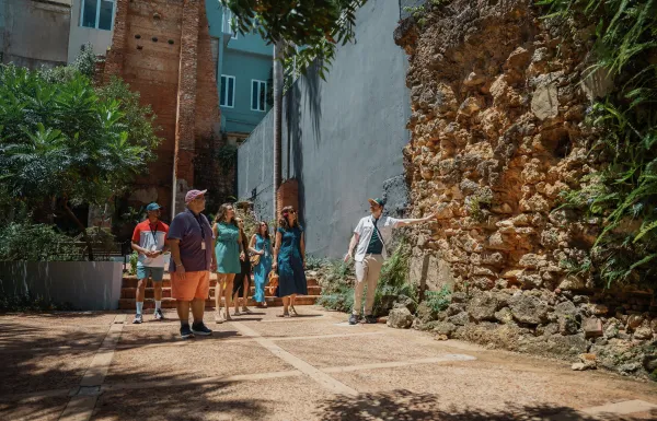 A tour guide points out historic stone ruins to a group visiting a courtyard in Old San Juan, Puerto Rico, surrounded by tropical greenery and colorful buildings.
