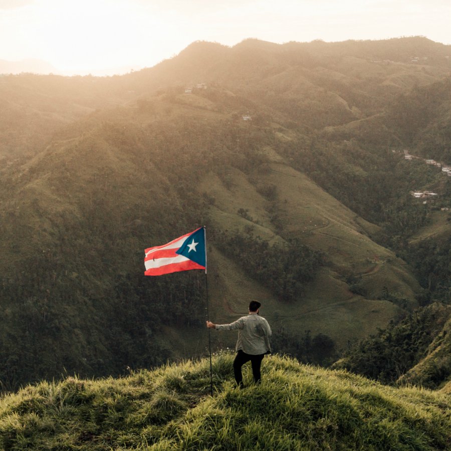 A man stands with a Puerto Rican flag atop a mountain summit as the sun sets.