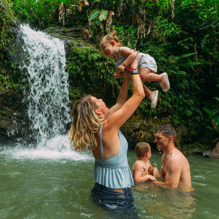 La madre sostiene a un bebé en el aire mientras está medio sumergida en una piscina de cascada del bosque.