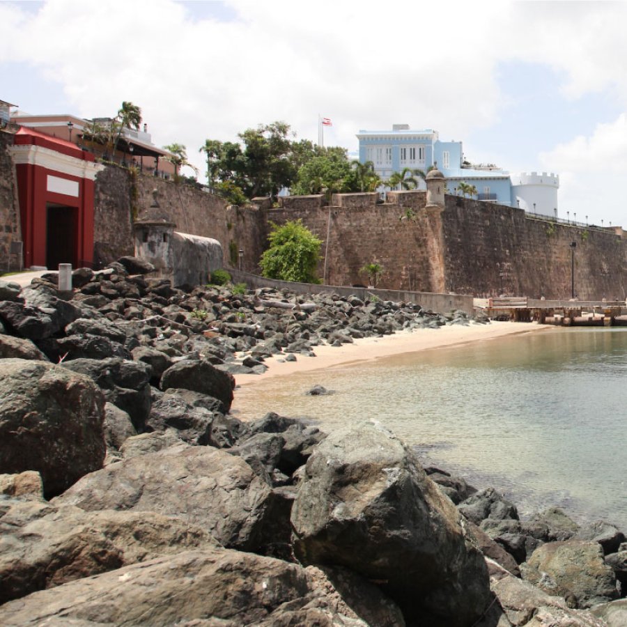 The bright-red gate of San Juan is pictured with rocks and water in the foreground.