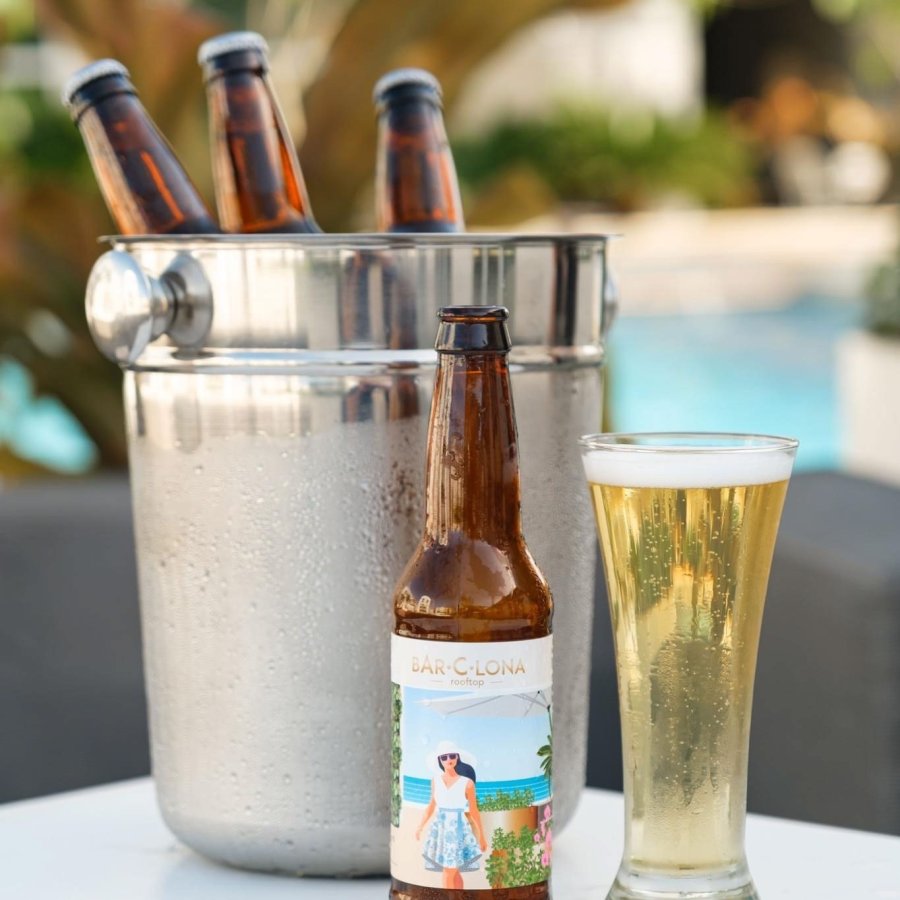 A bottle of Lola Bar-c-Lona beer stands next to a tall glass of beer and a silver bucket filled with several bottles of beer. AC Hotel by Marriott San Juan Condado.