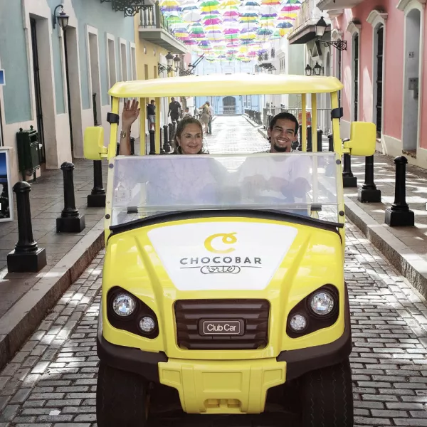 The "chocomobile," a yellow golf cart with Chocobar Cortes branding, carries a visitor through the streets of Old San Juan with colorful umbrellas overhead.