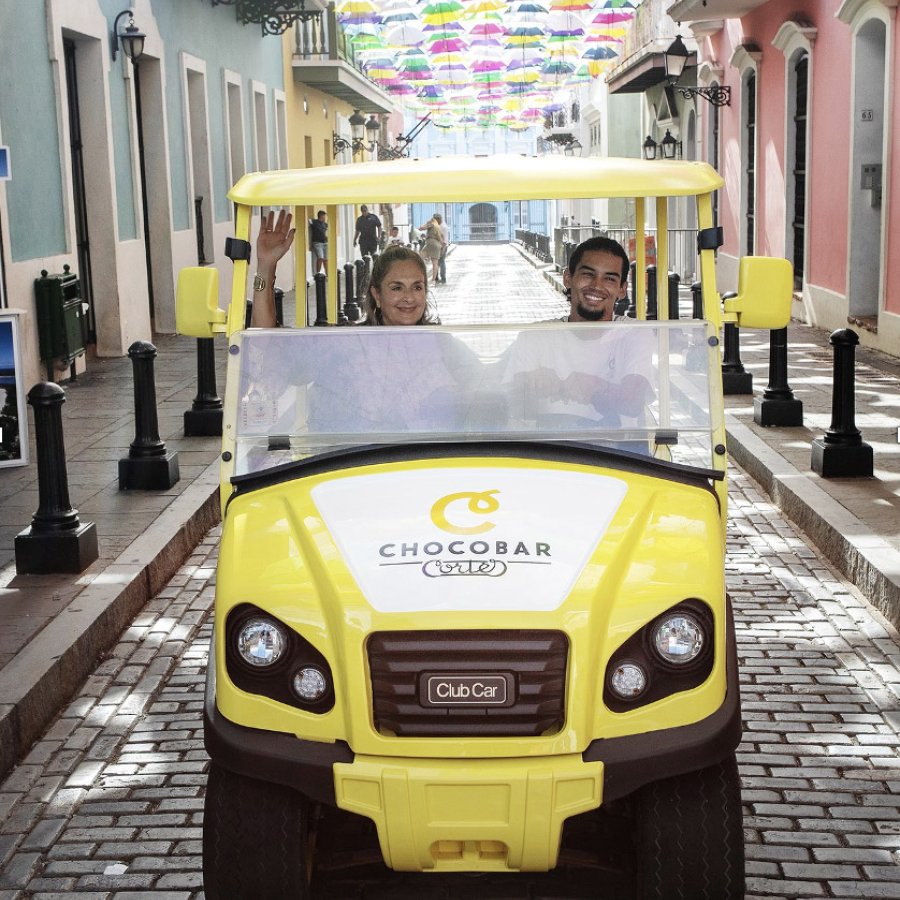 The "chocomobile," a yellow golf cart with Chocobar Cortes branding, carries a visitor through the streets of Old San Juan with colorful umbrellas overhead.