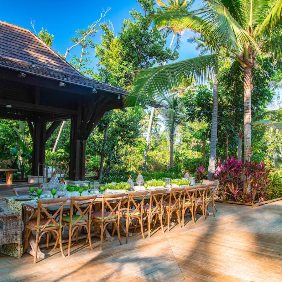 A long outdoor dining table with chairs, shaded by a pavilion and large palm trees, is set for dinner at St. Regis Bahia Beach Resort & Golf Club. 