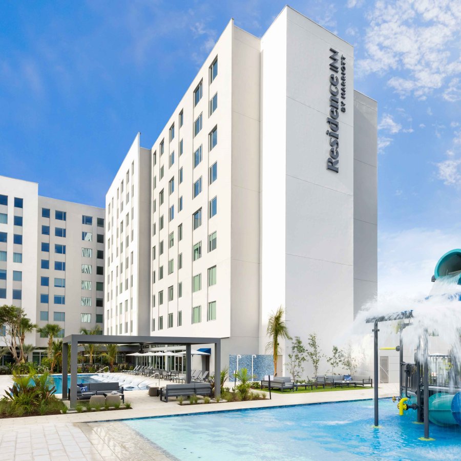 Photo showing a swimming pool with water fountain play area, with a tall white hotel building in the background. Residence Inn by Marriott San Juan Isla Verde.