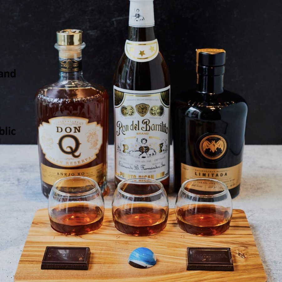 Rum and chocolate tasting presentation featuring three bottles of Puerto Rican rum, three glasses containing rum, and three chocolates on a wooden tray.