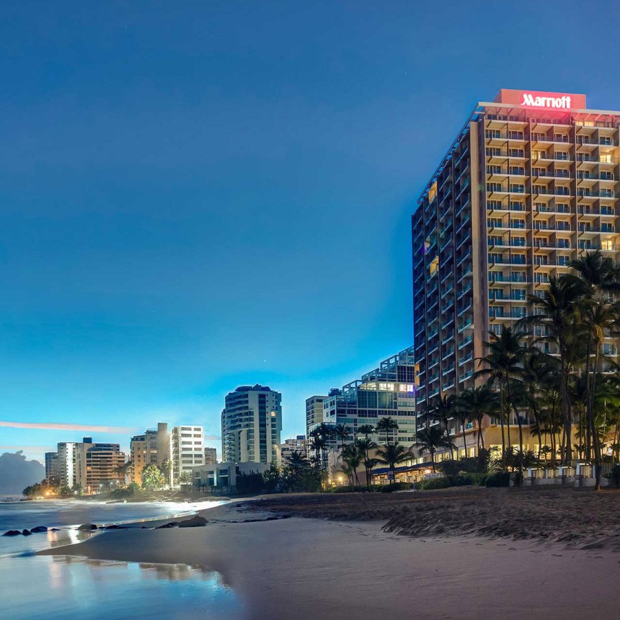 The exterior of the San Juan Marriott Resort & Stellaris Casino, with the word "Marriott" illuminated, with the beach in the foreground and the night sky behind it.