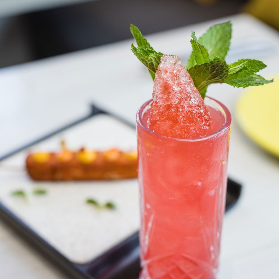 A bright pink cocktail with a green garnish is pictured on a table with a plate of food in the background.