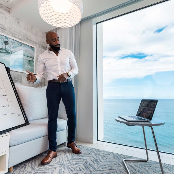 A man stands in front of a white board and a large window with an ocean view while making a remote presentation to his computer.