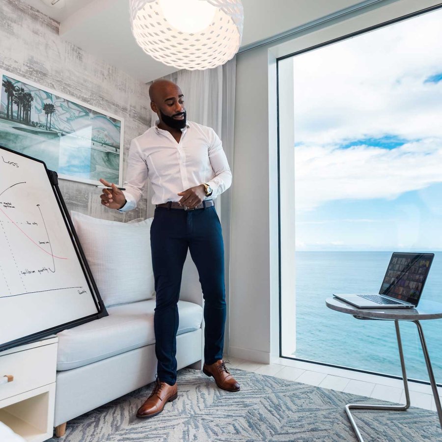 A man stands in front of a white board and a large window with an ocean view while making a remote presentation to his computer.