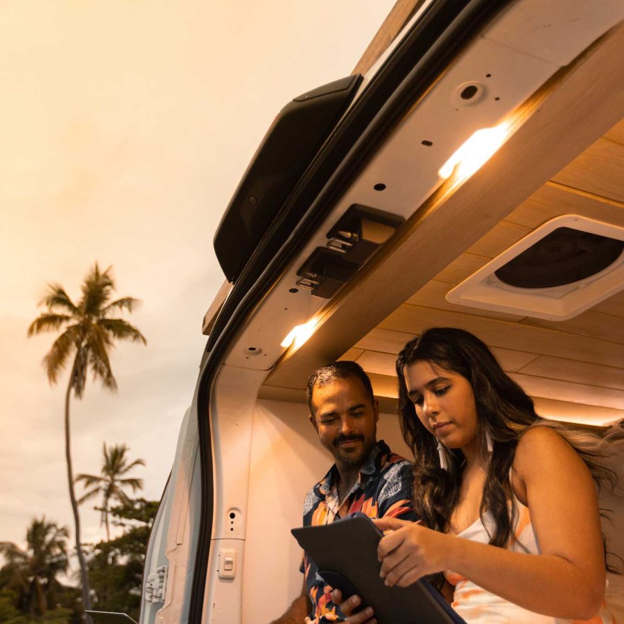 A couple works on tablets while sitting in the back of a van with the back open.