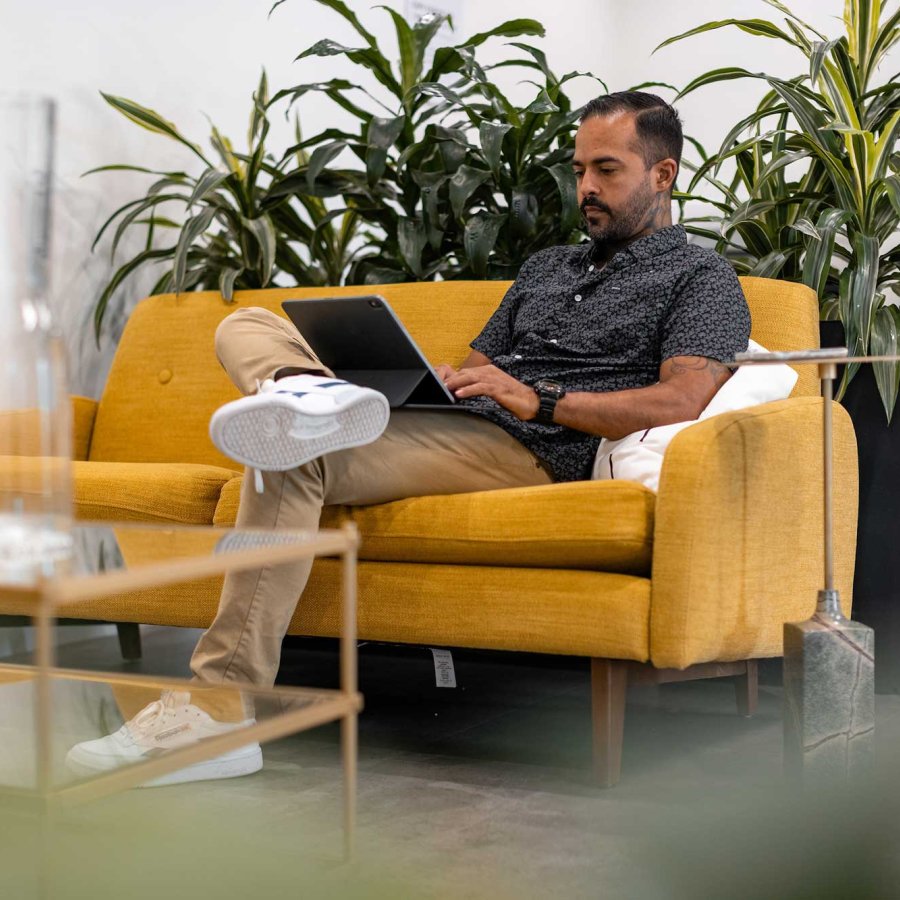 A man works on his laptop in a modern office while sitting on a yellow sofa.