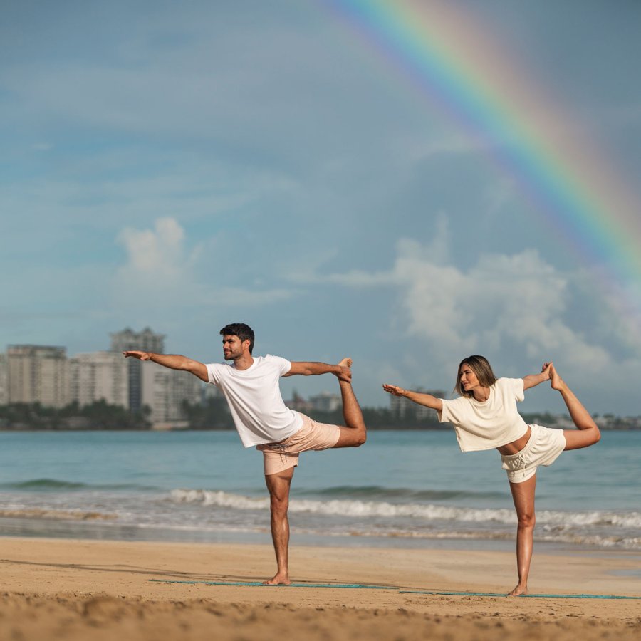 A couple practices yoga on a beach with a rainbow overhead at Fairmont El San Juan hotel.