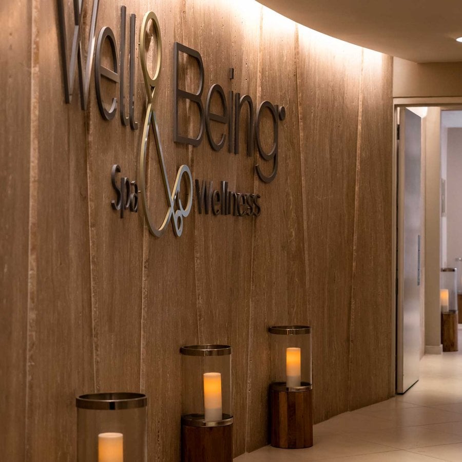 A man in a spa robe walks down a long hallway in front of a wooden wall with the words "Well & Being."
