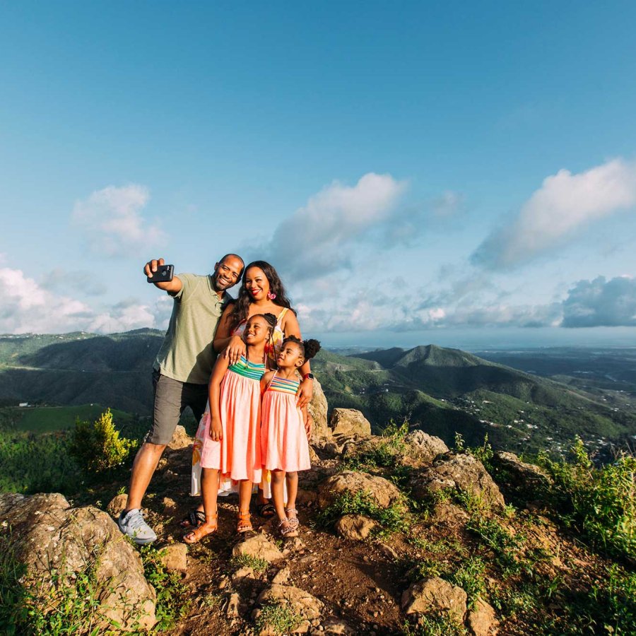 A mother, father, and two young girls pose for a selfie on top of a mountain with a dramatic backdrop in Puerto Rico.