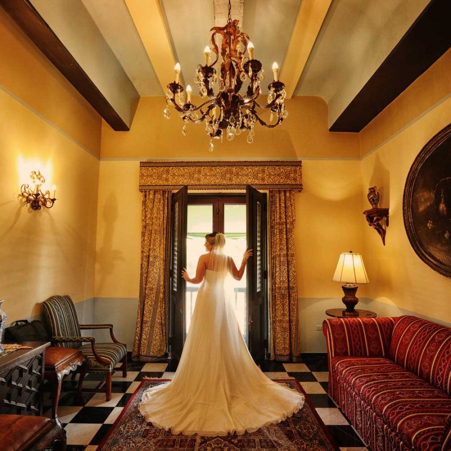 A bride, pictured from behind, opens a large historic window at Hotel El Convento in Old San Juan, Puerto Rico. Photo by Noel Pilar.