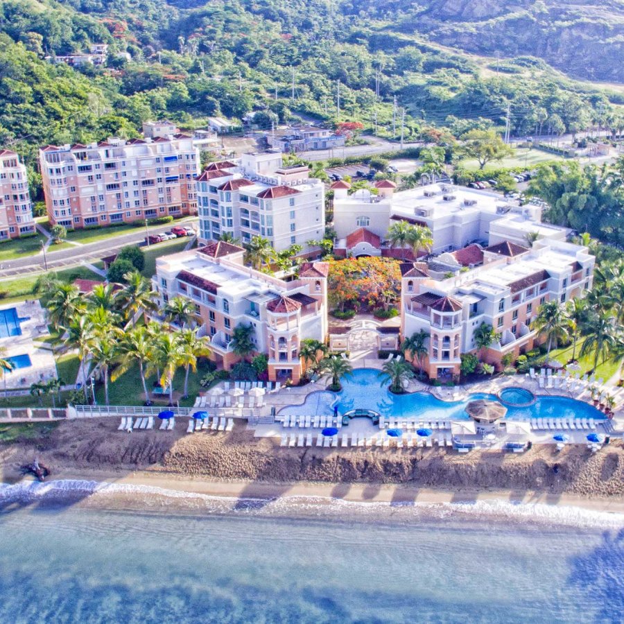 Overhead view of a large oceanfront resort with rolling green hills behind it. Rincon Beach Resort, Añasco, Puerto Rico.