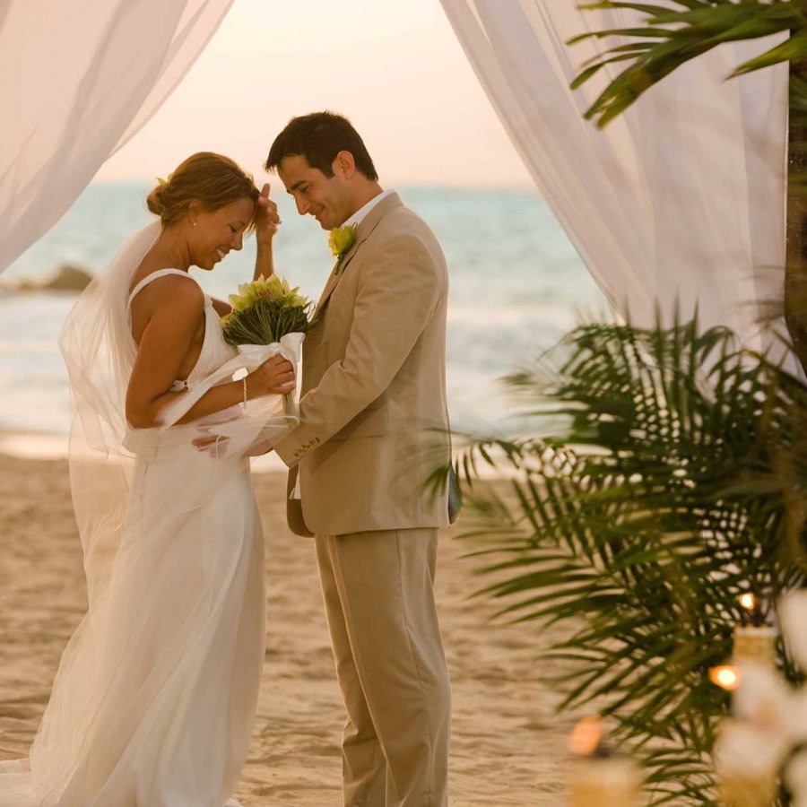A bride and groom face each other in front of an altar draped with white fabric on a beach in Puerto Rico.