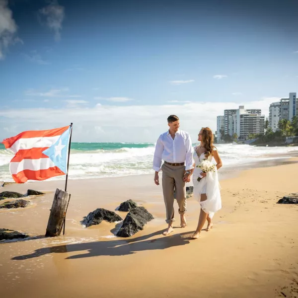 A bride and groom walk on the beach near the San Juan Marriott Resort & Stellaris Casino, with a Puerto Rican flag planted in the sand.