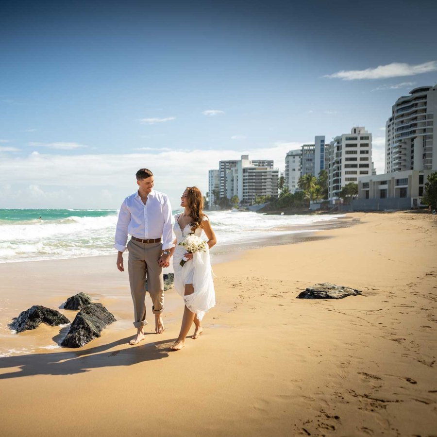 A bride and groom walk on the beach near the San Juan Marriott Resort & Stellaris Casino, with a Puerto Rican flag planted in the sand.
