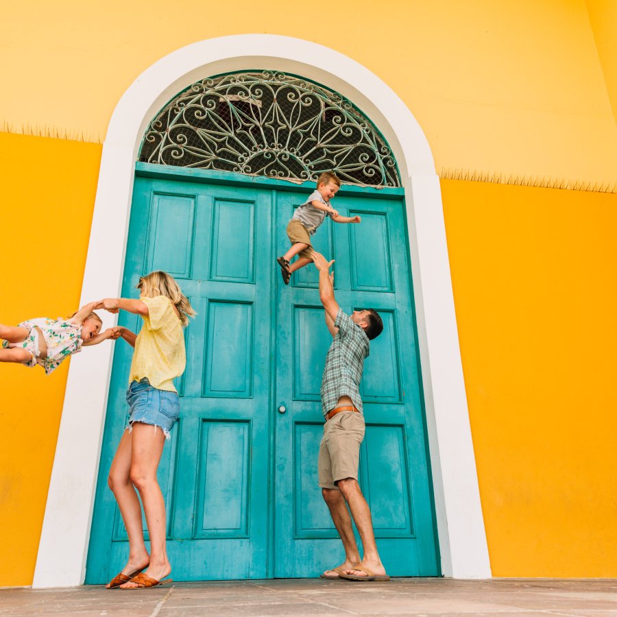 A couple plays with their children. The family stands next to a vivid yellow wall with a teal blue door.