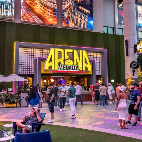 Colorful neon sign for Arena Medalla, a sports bar at Distrito T-Mobile in San Juan, Puerto Rico. People gather in front of the building.