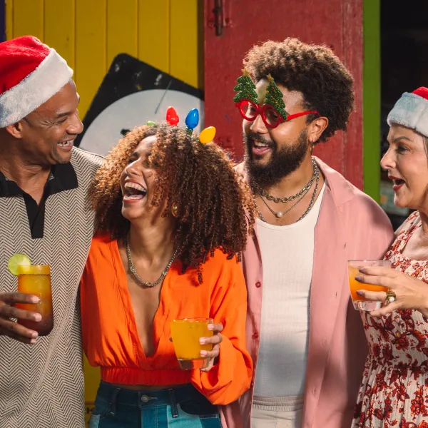 Four people laugh together in Santa hats and festive accessories, with three holding tropical drinks.