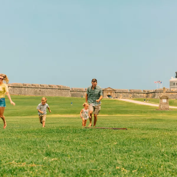 A family enjoys a sunny day at El Morro in San Juan, Puerto Rico, with children running across the green lawn.