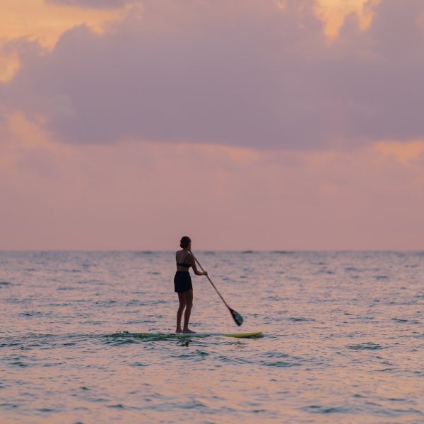 A woman paddleboards alone on calm ocean waters under a pastel-colored sunset sky, creating a peaceful and serene scene.