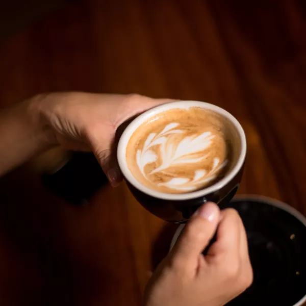 A person holding a black cup of coffee with latte art on top, over a wooden table.