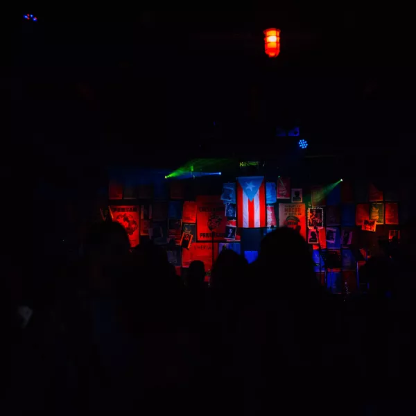 Dark stage with a Puerto Rican flag, colorful lights, and a crowd watching.