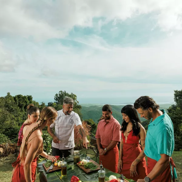 Group gathered around a chef preparing fresh local ingredients on a table covered with banana leaves, overlooking lush mountains and coastline in Puerto Rico.