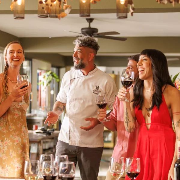 Chef leading a lively wine tasting indoors, as guests laugh and raise glasses around a table set with charcuterie and fresh ingredients.