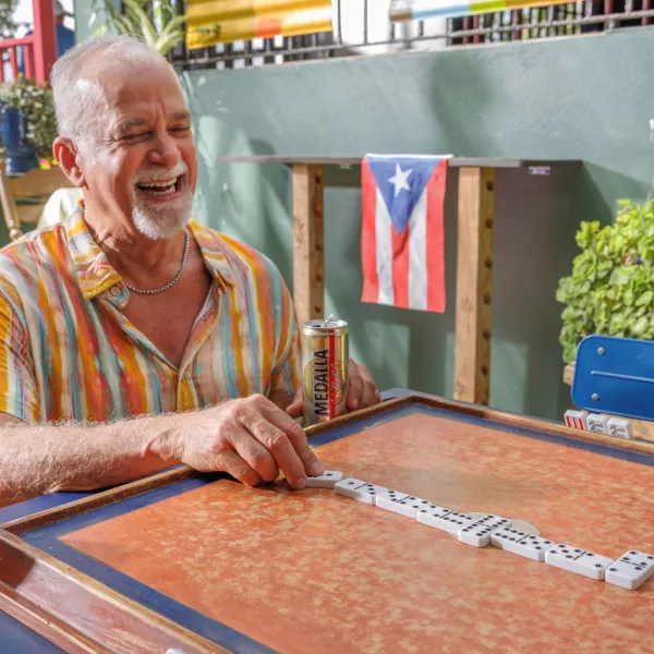Older man laughing while playing dominoes at an outdoor table in Puerto Rico, with a Puerto Rican flag hanging in the background.