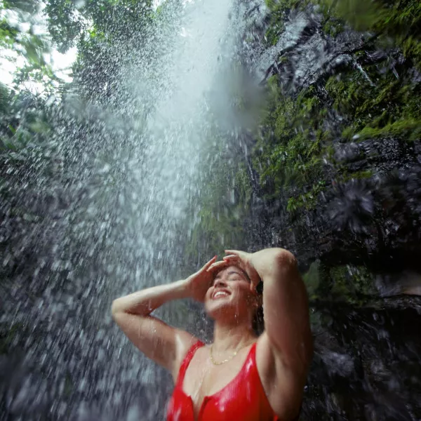 Woman in a red swimsuit standing beneath a waterfall in El Yunque National Forest, Puerto Rico, smiling with eyes closed as water cascades around her in the lush tropical rainforest.