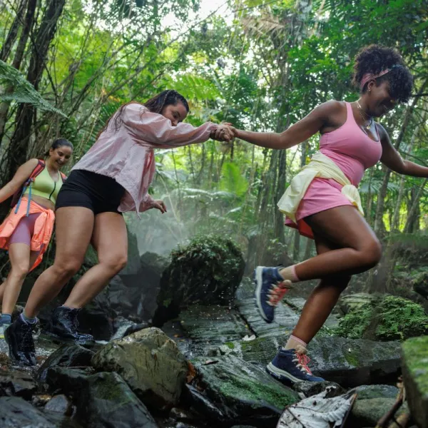 Three women hiking and carefully crossing rocks in a lush tropical rainforest in Puerto Rico, surrounded by dense greenery and mist.