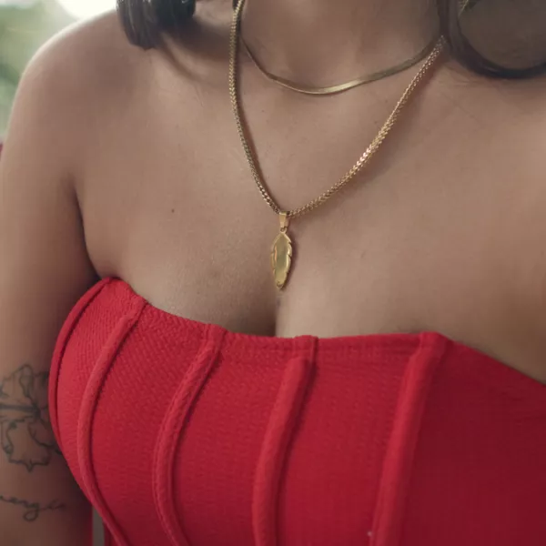 Close-up of a woman wearing a red strapless top, adorned with gold jewelry from local brand, El Papelón. Tattoos are visible on her arm, with a blurred outdoor background.
