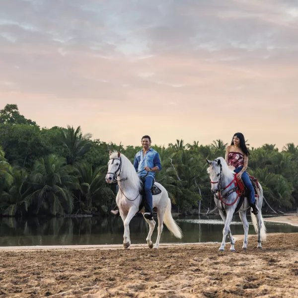 Two riders on white horses walk along a sandy beach beside a calm river in Luquillo, surrounded by lush greenery and a colorful sky.