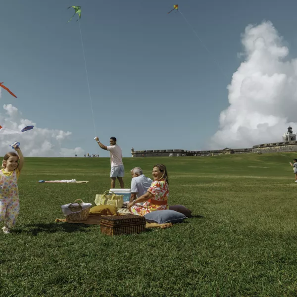 A family enjoys a picnic on a grassy hill, flying colorful kites from the local brand Chiringas.com under a blue sky with fluffy clouds. The historic structure of El Morro is visible in the background.