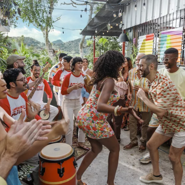 A vibrant outdoor gathering features diverse people dancing and playing instruments, surrounded by lush greenery and colorful decor.