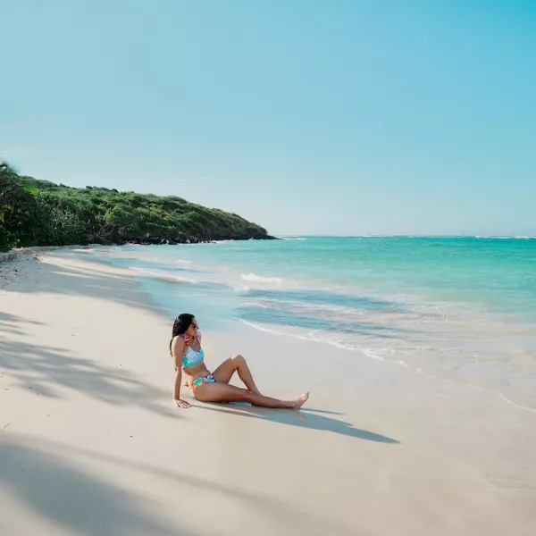 Woman sitting on the white sand at Flamenco Beach in Culebra, looking out at the calm turquoise water under a clear Caribbean sky.