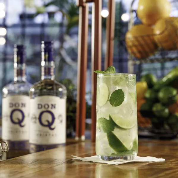 A refreshing mojito with mint and lime sits on a wooden bar, with two bottles of Don Q rum in the background and a fruit basket nearby.