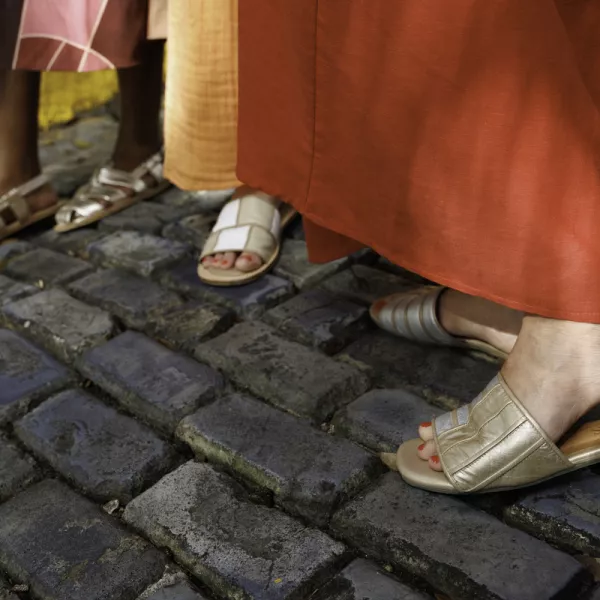 Three pairs of feet in stylish sandals from local brand Isleña stand on a cobblestone street, with vibrant clothing flowing around them. 