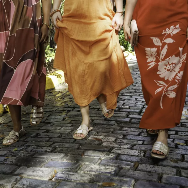 Three women walk along a cobblestone path, wearing colorful dresses and sandals from local brand Isleña. 