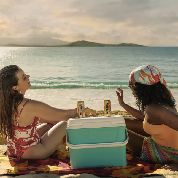 Two women sit on a sandy beach, facing each other, with a cooler and beers between them. The ocean sparkles in the background under a cloudy sky.