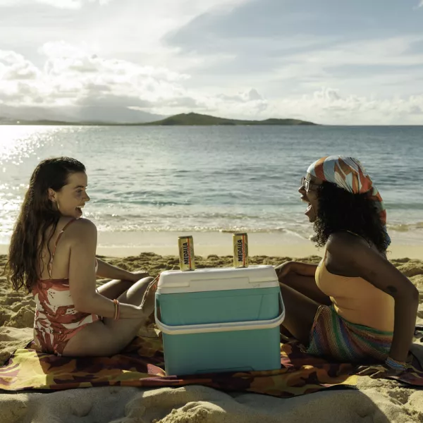 Two women sit on a sandy beach, facing each other with a cooler and beers between them. The ocean sparkles in the background under a bright sky.