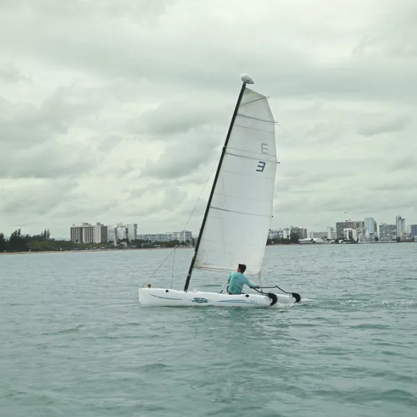 A small sailboat glides across calm waters under a cloudy sky, with Isla Verde visible in the background. 