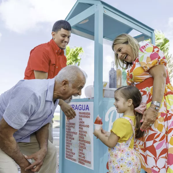 A colorful piraguas stand with a family enjoying treats outdoors. A child in a yellow shirt interacts with an elderly man, while adults smile.