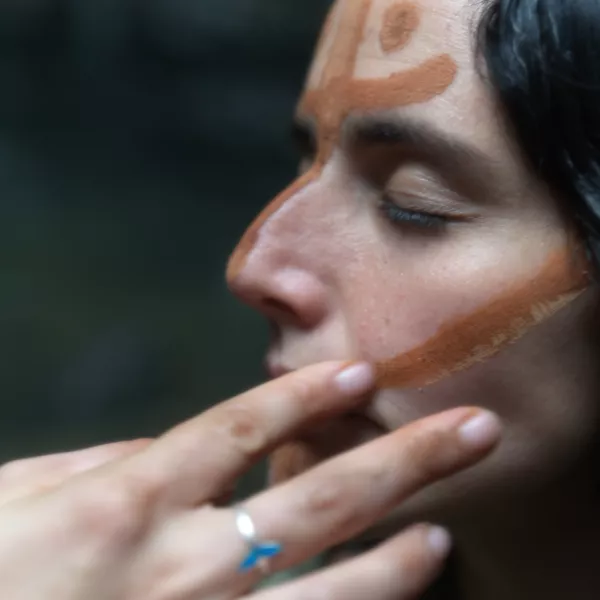 Close-up of a woman with eyes closed as another person gently applies earthy-toned face paint across her cheek and forehead.