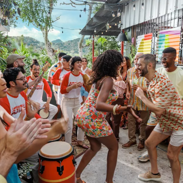 A lively outdoor gathering with people dancing to live music, playing drums and guitar, and celebrating together on a colorful patio surrounded by tropical plants and mountain views.